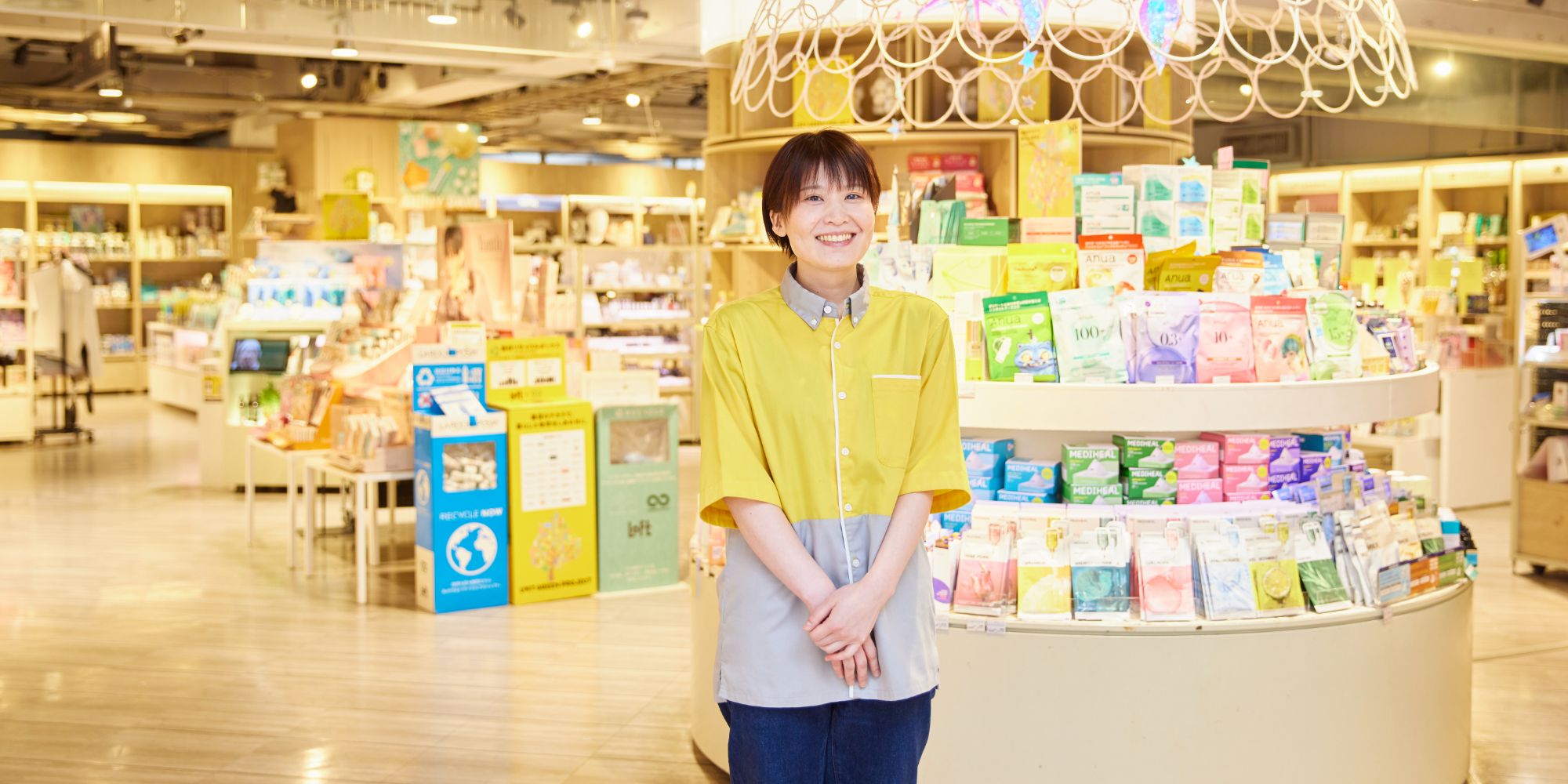 化粧品の円形ディスプレイの前に笑顔で立つS.E.さん