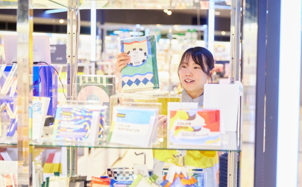 カラフルな文具の品出しをしているT.Y.さんがガラス棚越しに写っている様子